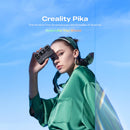 A woman in a green satin blouse holds a black Creality Pika portable AI scanner against her face, set against a bright blue sky background with promotional text.