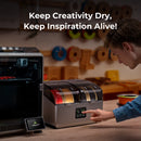 Person interacting with a 3D printing filament dryer with text 'Keep Creativity Dry, Keep Inspiration Alive!' in a workshop setting.